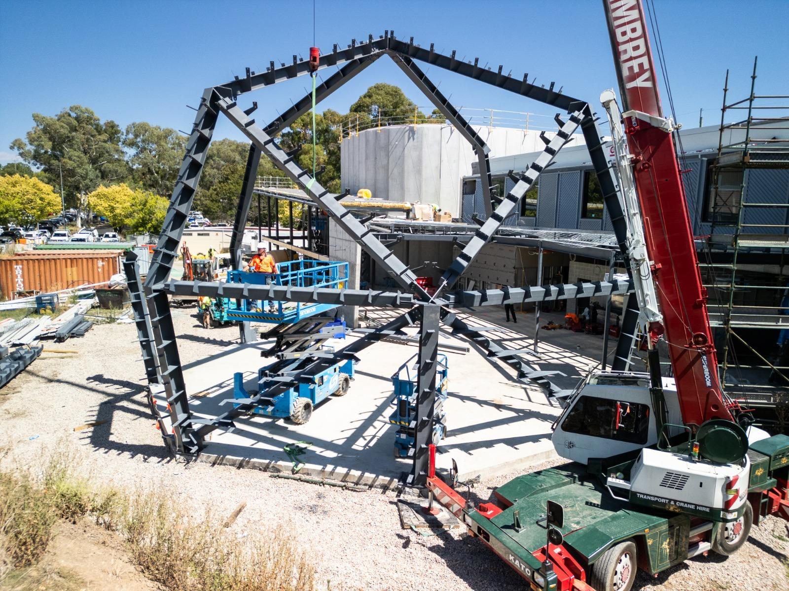 Large painted steel feature frame being erected on site with a city crane and scissor lifts